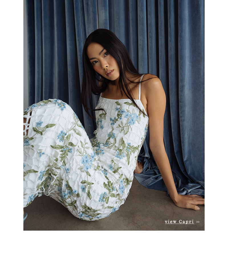A woman in a white dress with blue floral patterns sits on a concrete floor, leaning against blue velvet curtains. She appears relaxed and contemplative.