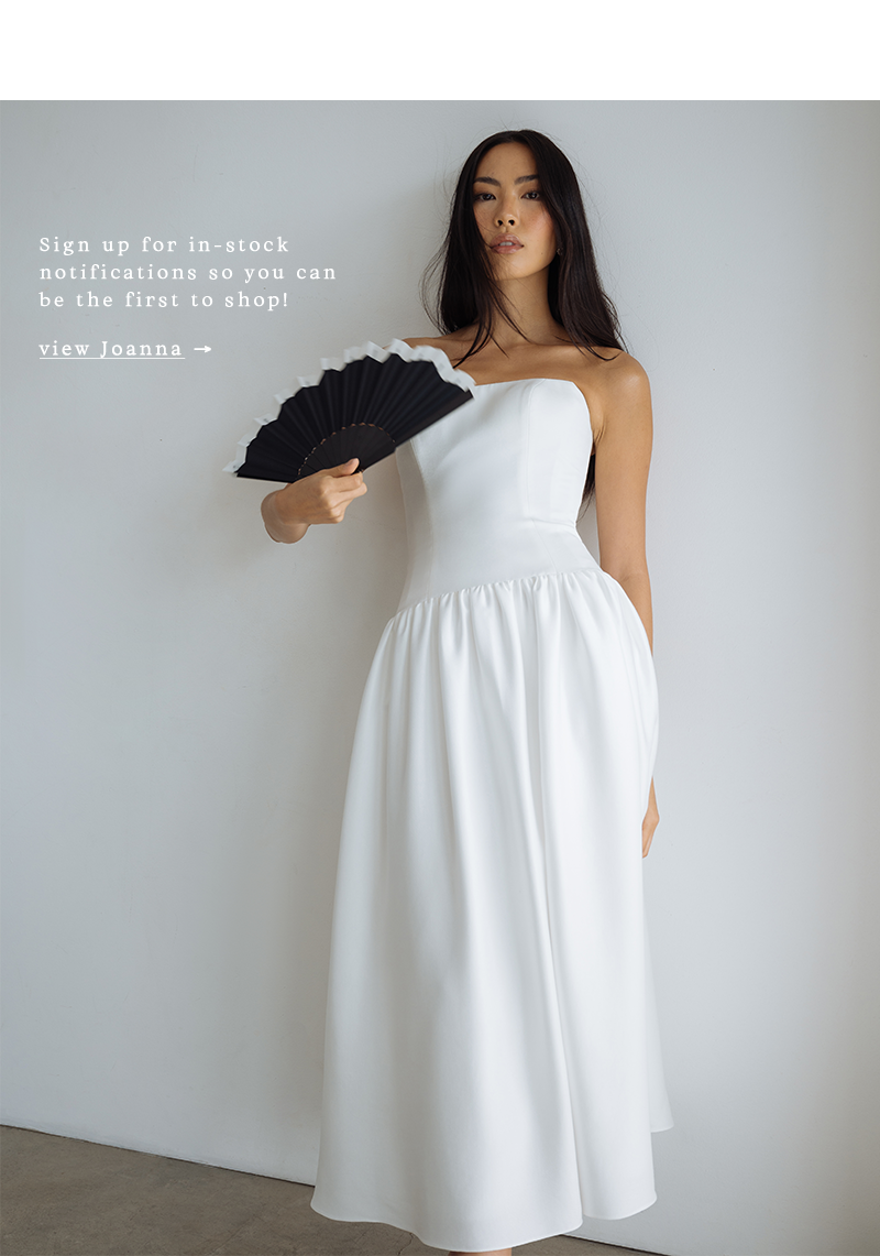 A woman in a strapless white dress stands against a plain wall, holding a black fan. The text promotes signing up for in-stock notifications. Elegant and poised tone.