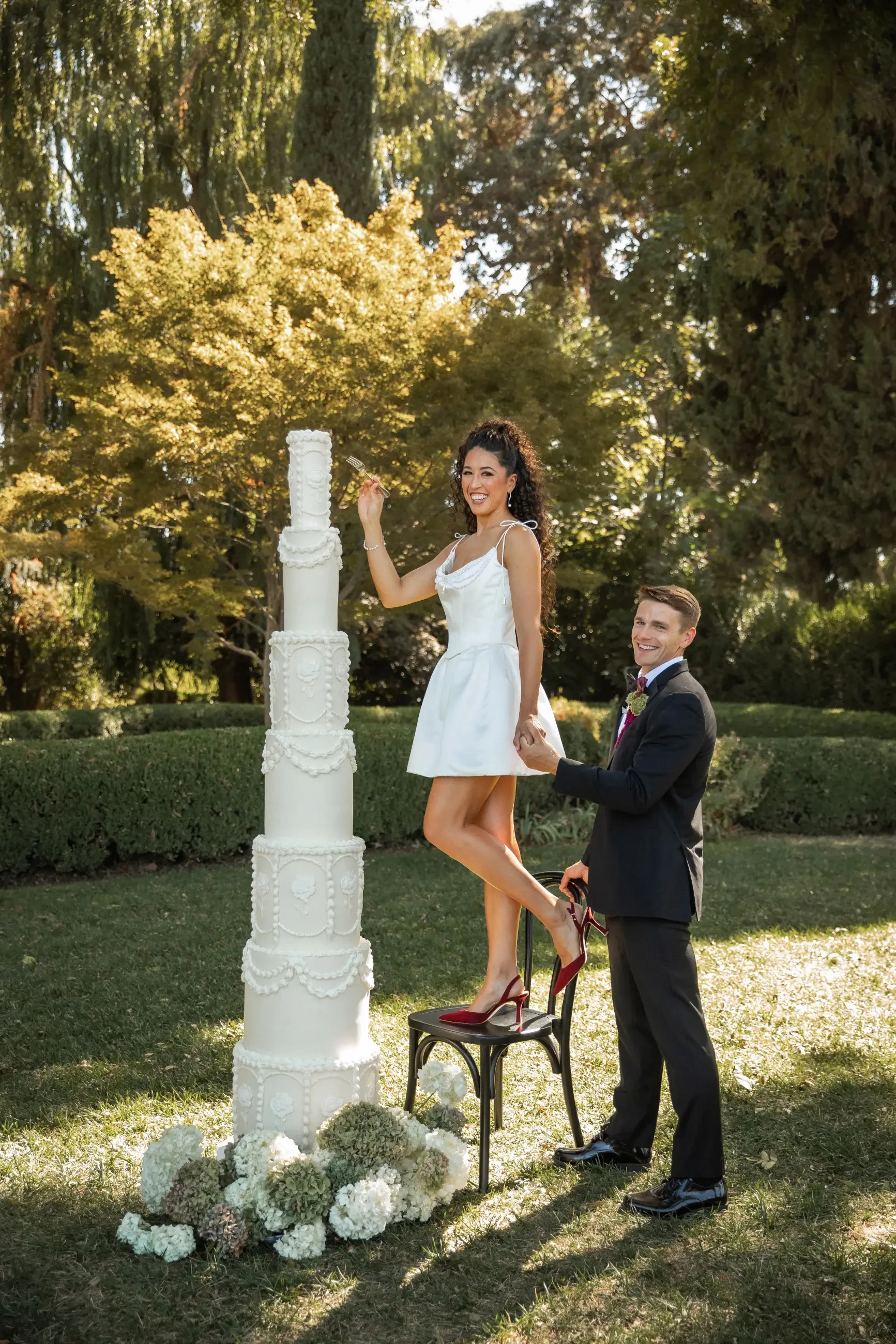 Newlyweds smiling outdoors, with the bride in a short white dress standing on a chair beside a towering, ornate wedding cake. The groom holds her hand, indicating joy and celebration in a lush garden setting.