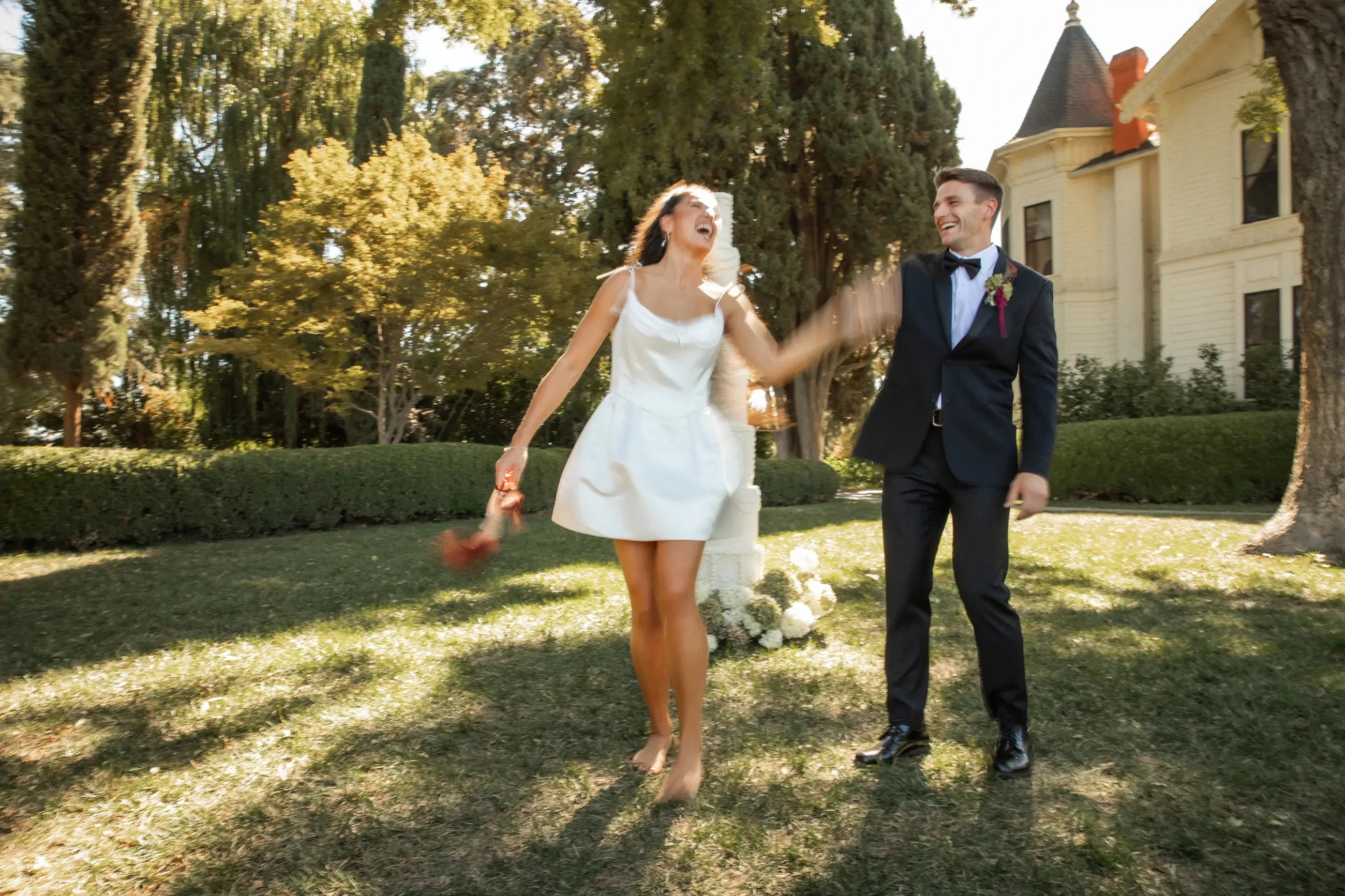 A joyful couple dances on a sunny lawn, the woman in a white dress and the man in a suit. A historic building and lush trees create a warm backdrop.
