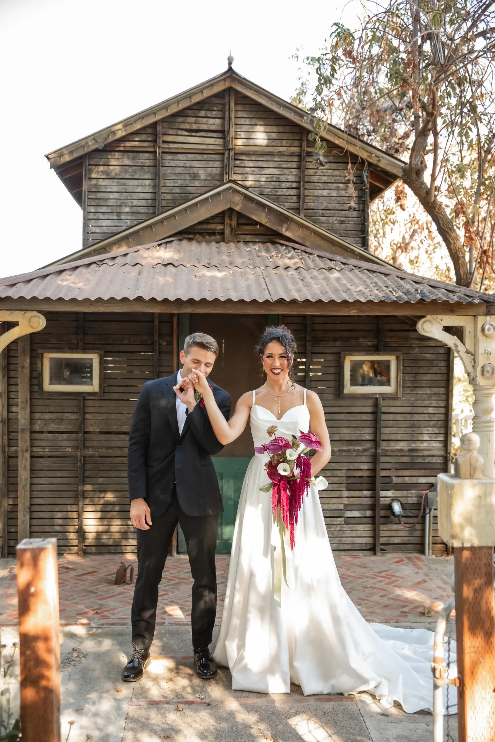 A bride in a white gown holds a bouquet, smiling as a groom in a black suit kisses her hand. They stand in front of a rustic wooden cabin.