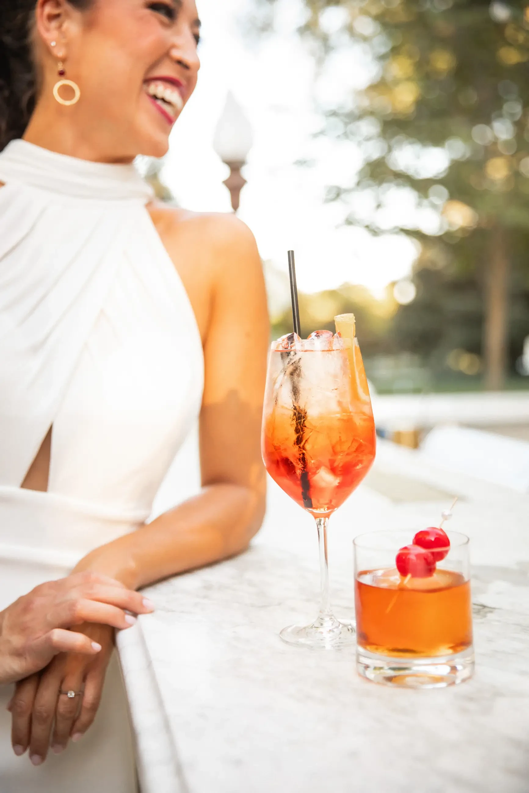 A woman in a white dress smiles near a marble counter with two cocktails: a vibrant orange drink with ice and a glass with two red cherries. The mood is cheerful.