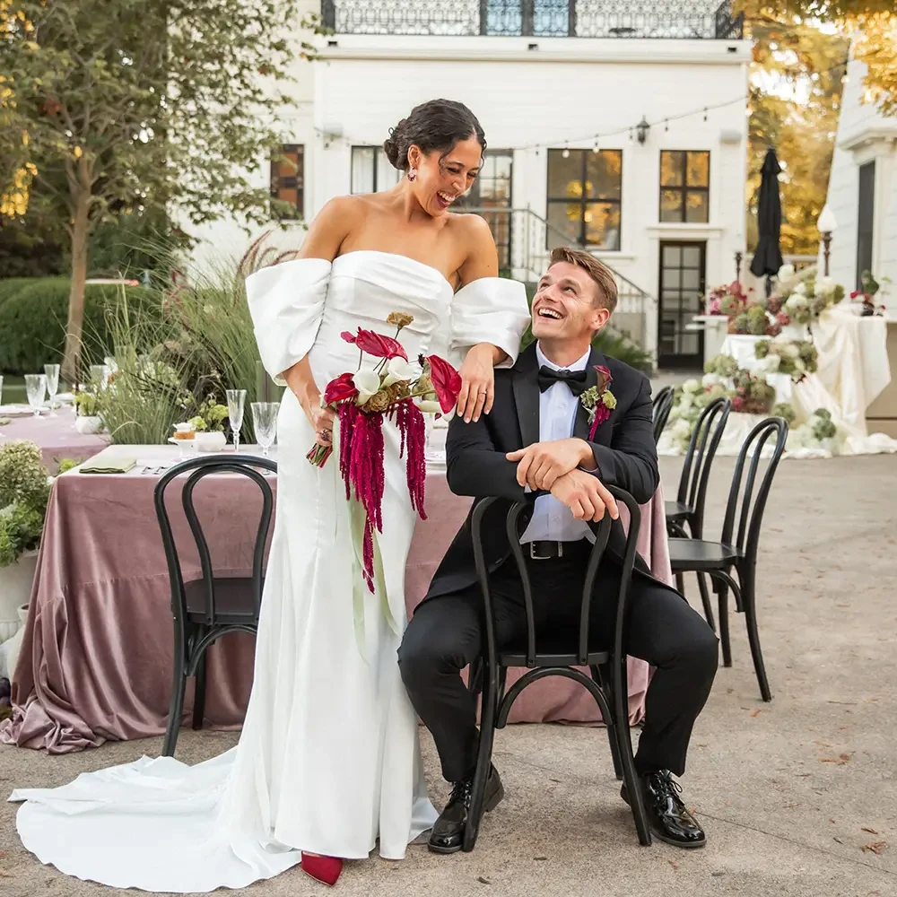 Bride in white gown and groom in black suit share a joyful moment outdoors. She holds a bouquet and leans on him, surrounded by elegant decor.