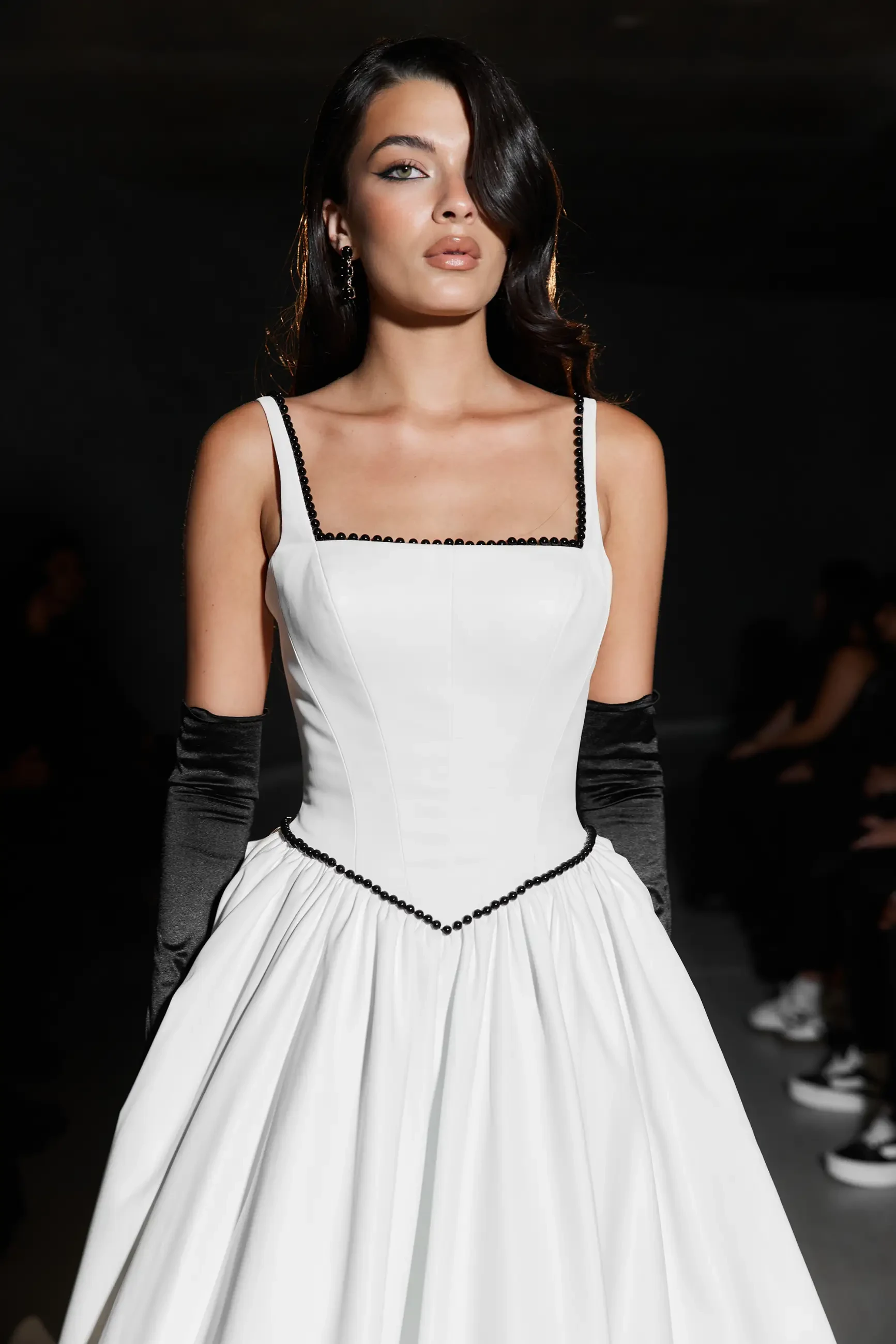 Model is wearing a wedding dress featuring a modern square neckline, a Basque waist with black-beaded bodice accents, and a full A-line skirt finished with a dramatic bow and buttoned train.