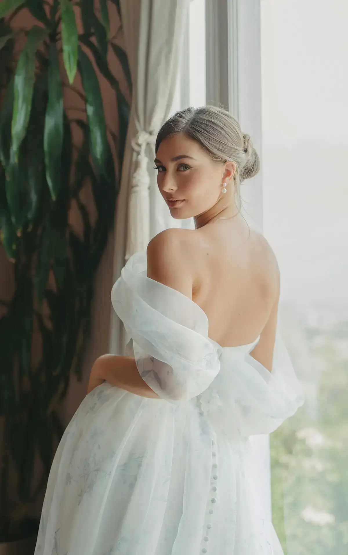 A woman in a backless white dress with sheer sleeves is standing near a window, looking over her shoulder. There are green plants in the background.
