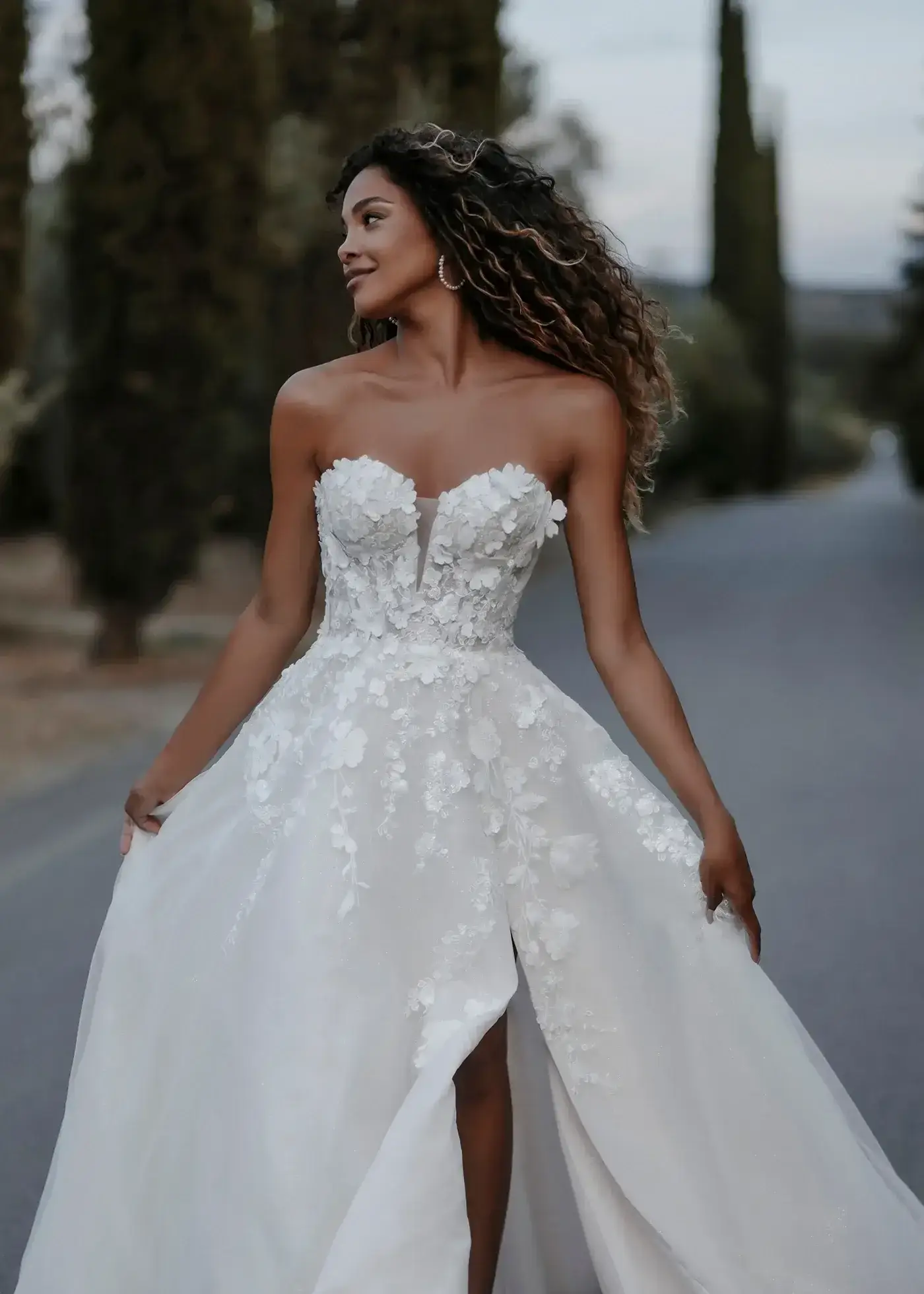 A woman wearing a strapless white wedding dress with floral details, walking along a road lined with trees, smiling and looking to the side.