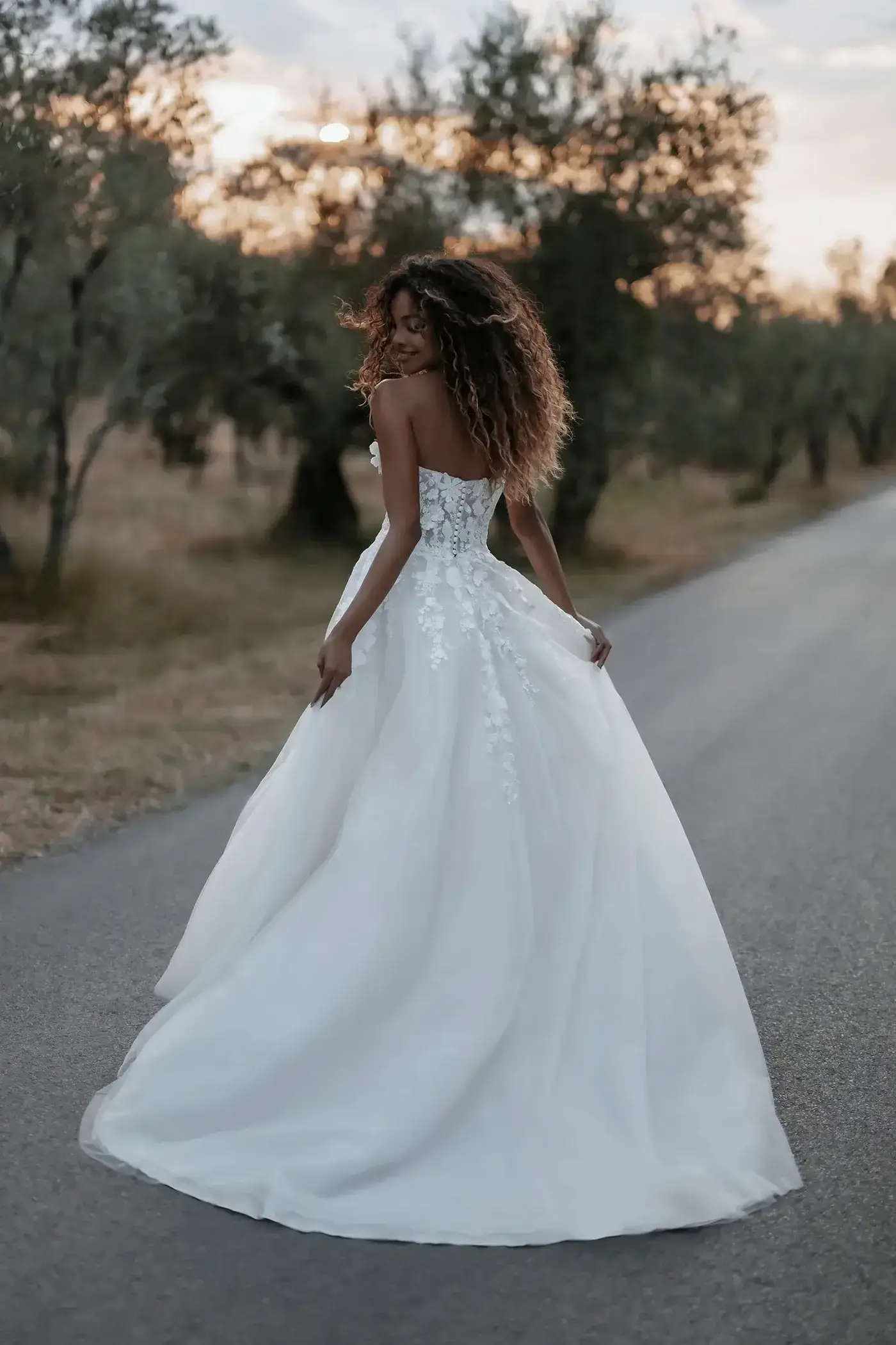 A woman in a long white gown stands on a road, facing away from the camera, with curly hair and trees in the background.