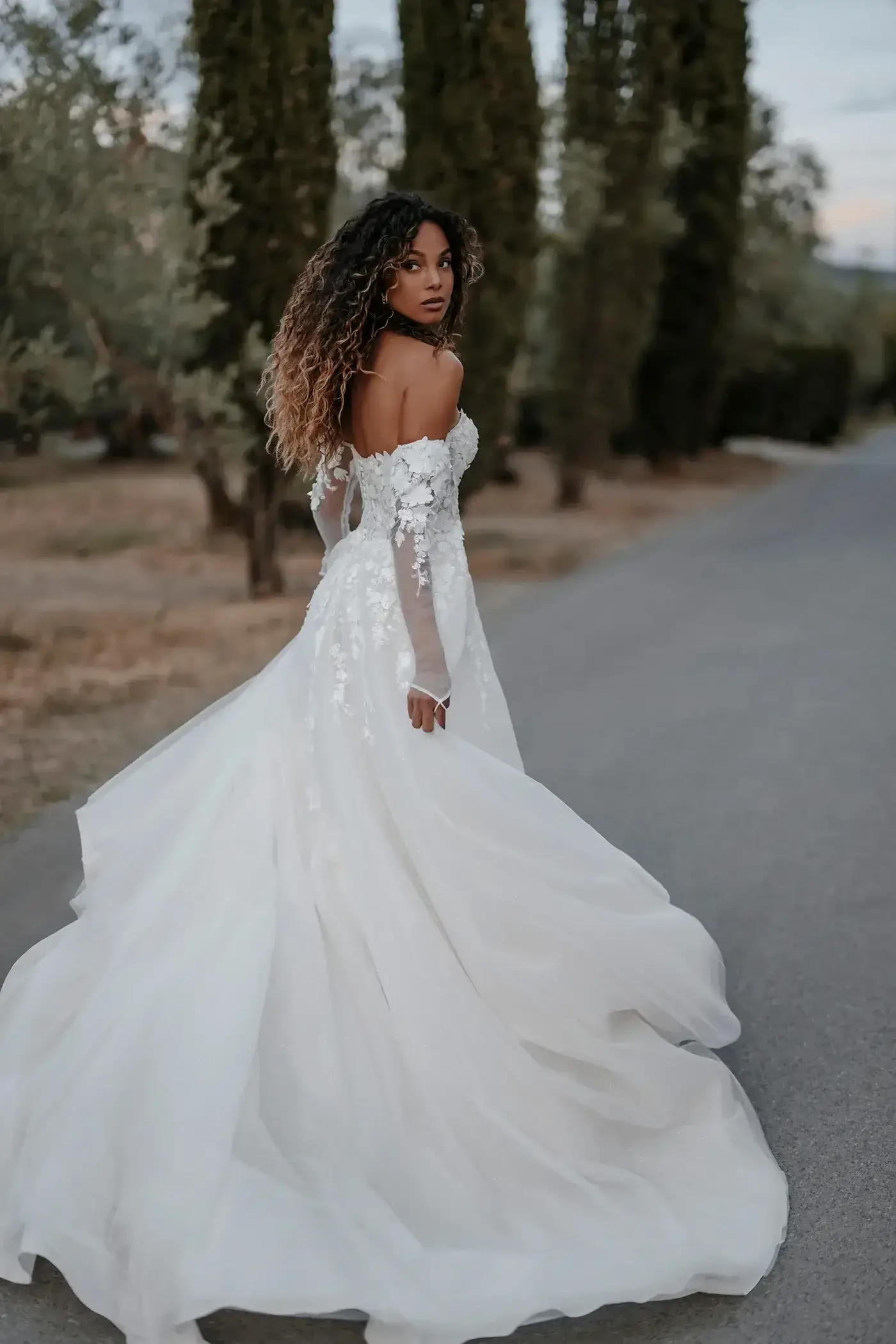 A bride in a flowing white wedding dress is walking away, looking back over her shoulder. The dress features off-the-shoulder sleeves and lace details. She has long, curly hair and is surrounded by tall trees along an empty road.