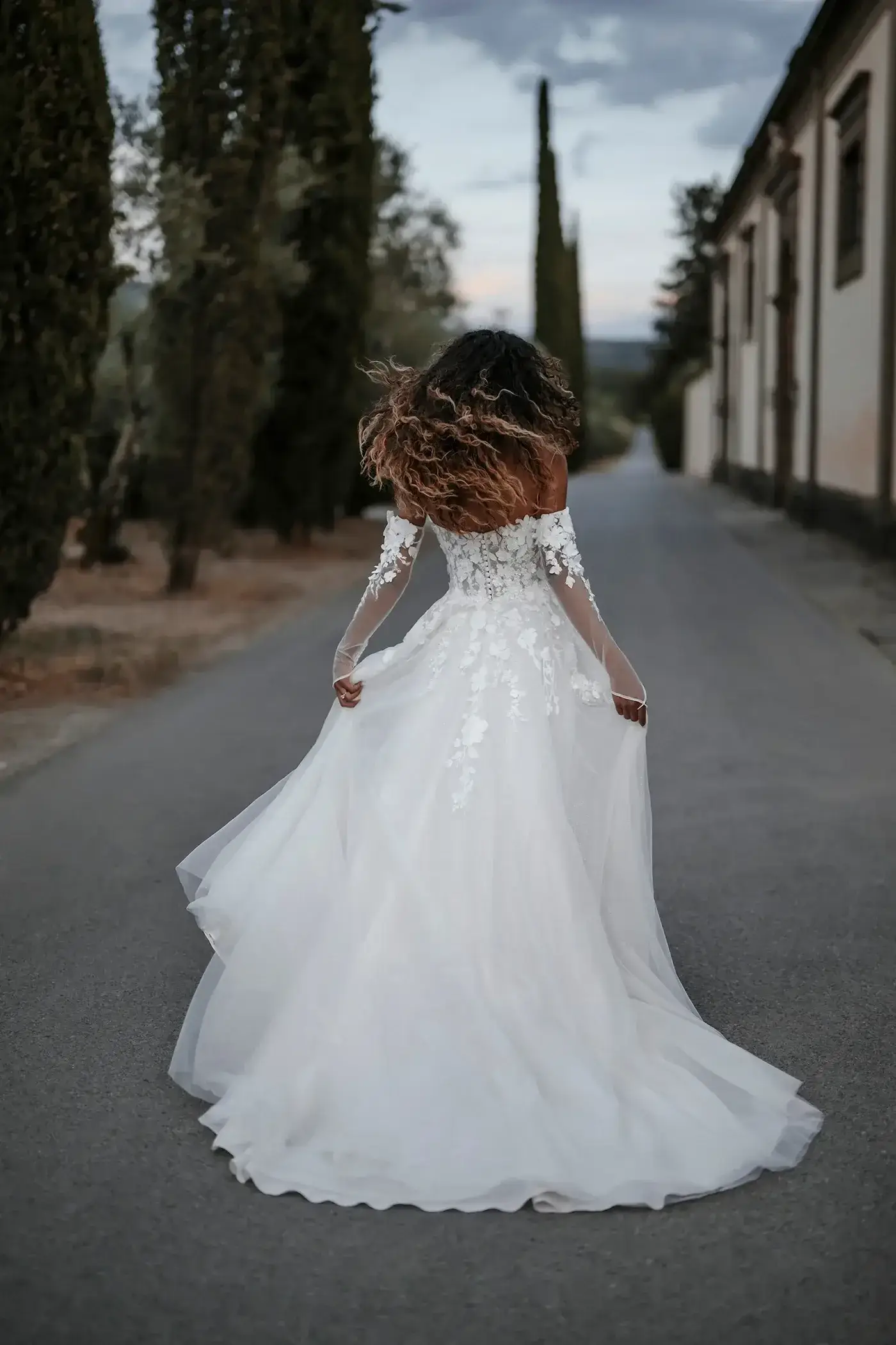 A woman in a flowing white wedding dress stands on a road, with her back to the camera, her hair blowing slightly in the breeze. Trees line the sides of the road leading towards buildings in the background under a cloudy sky.