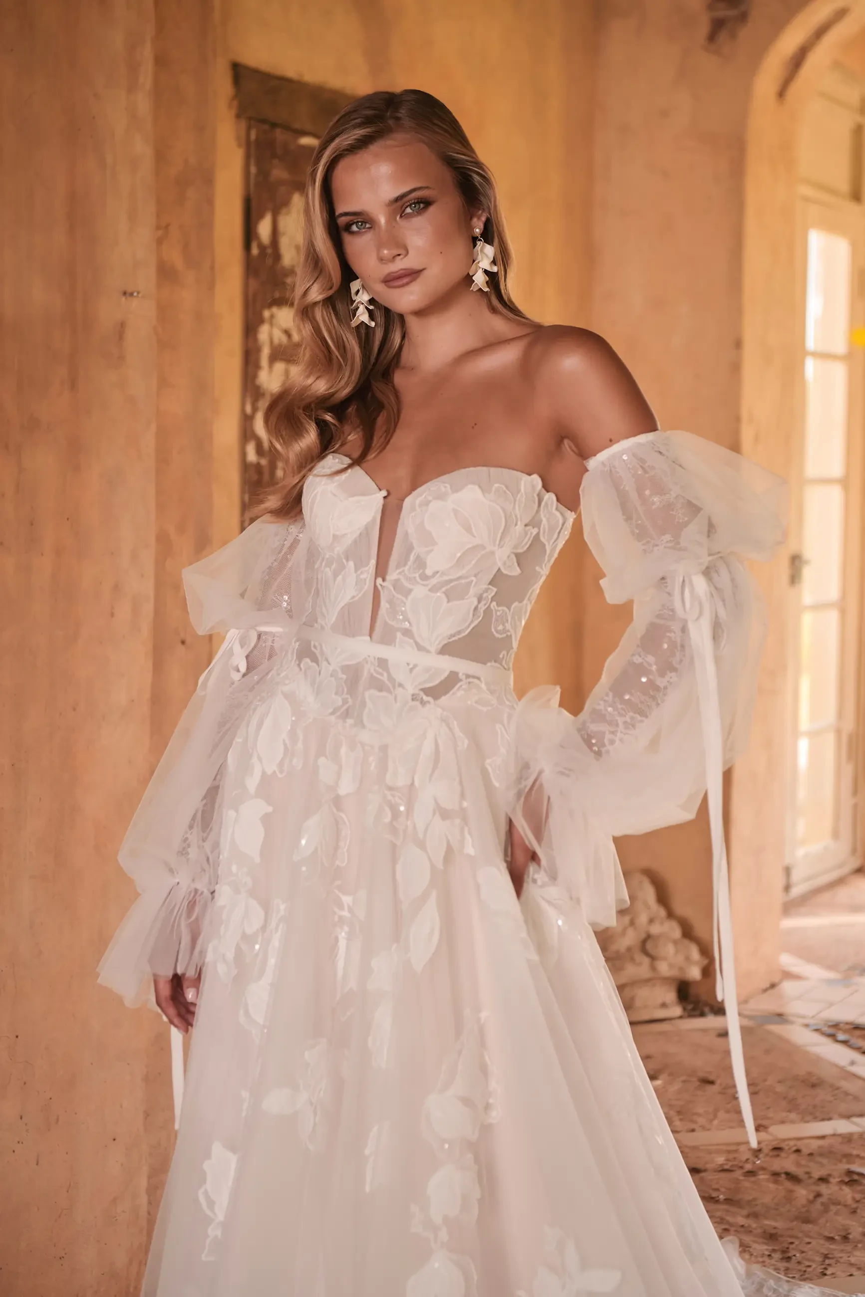 Model is wearing a wedding dress in layered tulle and organza with abstract floral lace, a Basque waist, micro-plunge bodice, and detachable blouson sleeves.