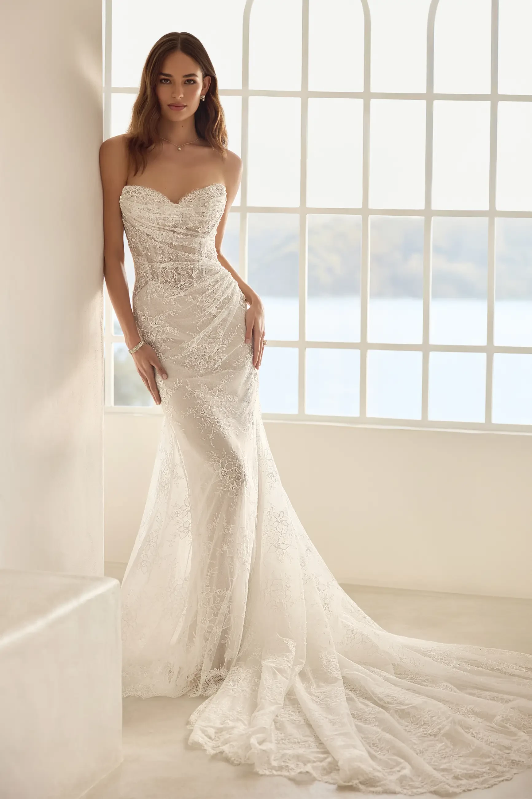A woman wearing a strapless, fitted white wedding dress stands by a large window with a soft natural light illuminating the scene.