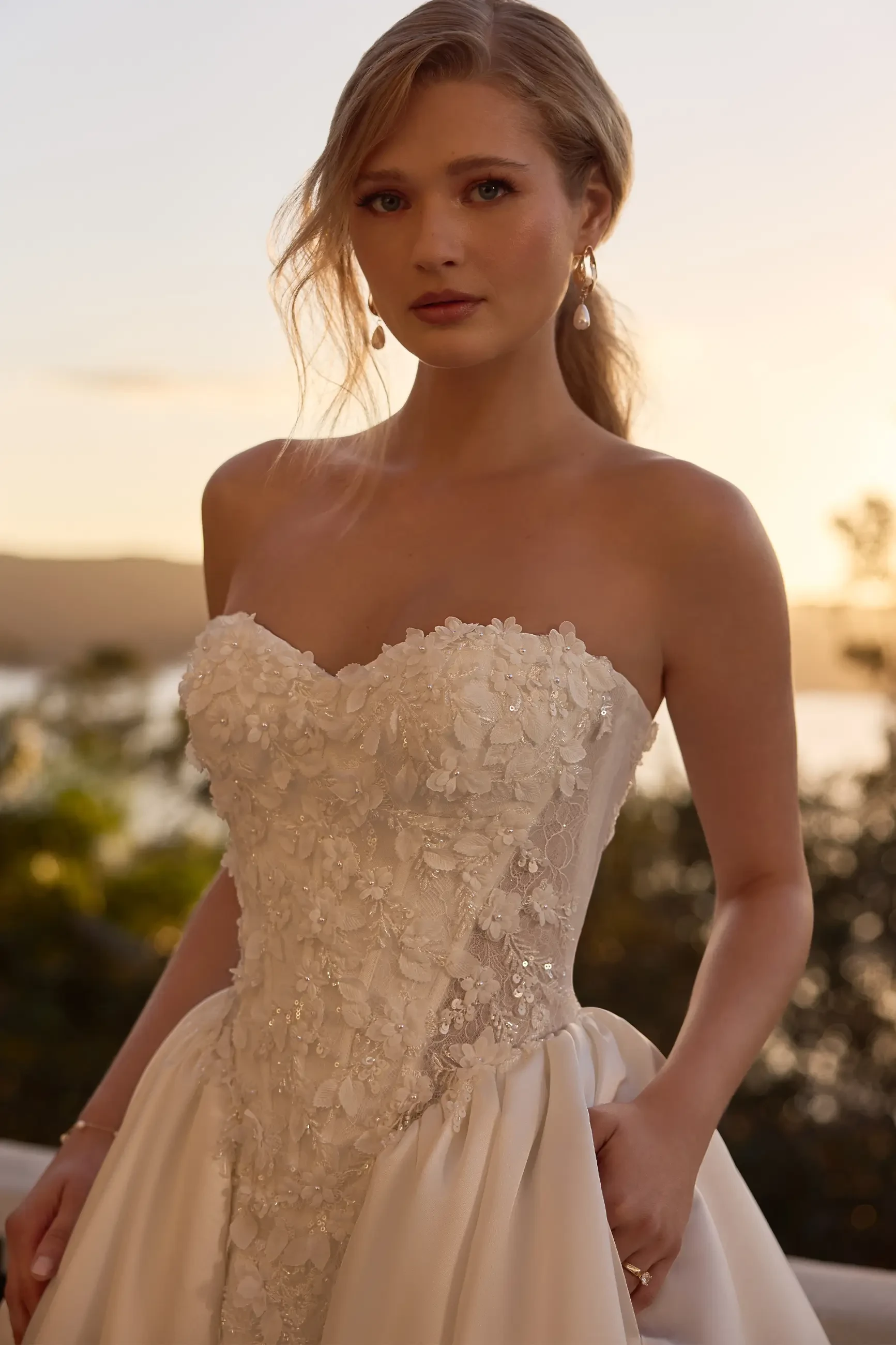 A woman in a strapless, floral-embellished white dress stands outdoors, with a soft sunset background. She has long hair pulled back and wears earrings.